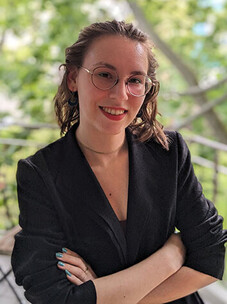 Frau Werle mit Brille, schwarzem Blazer und rotem Lippenstift, draußen vor grüner Kulisse.